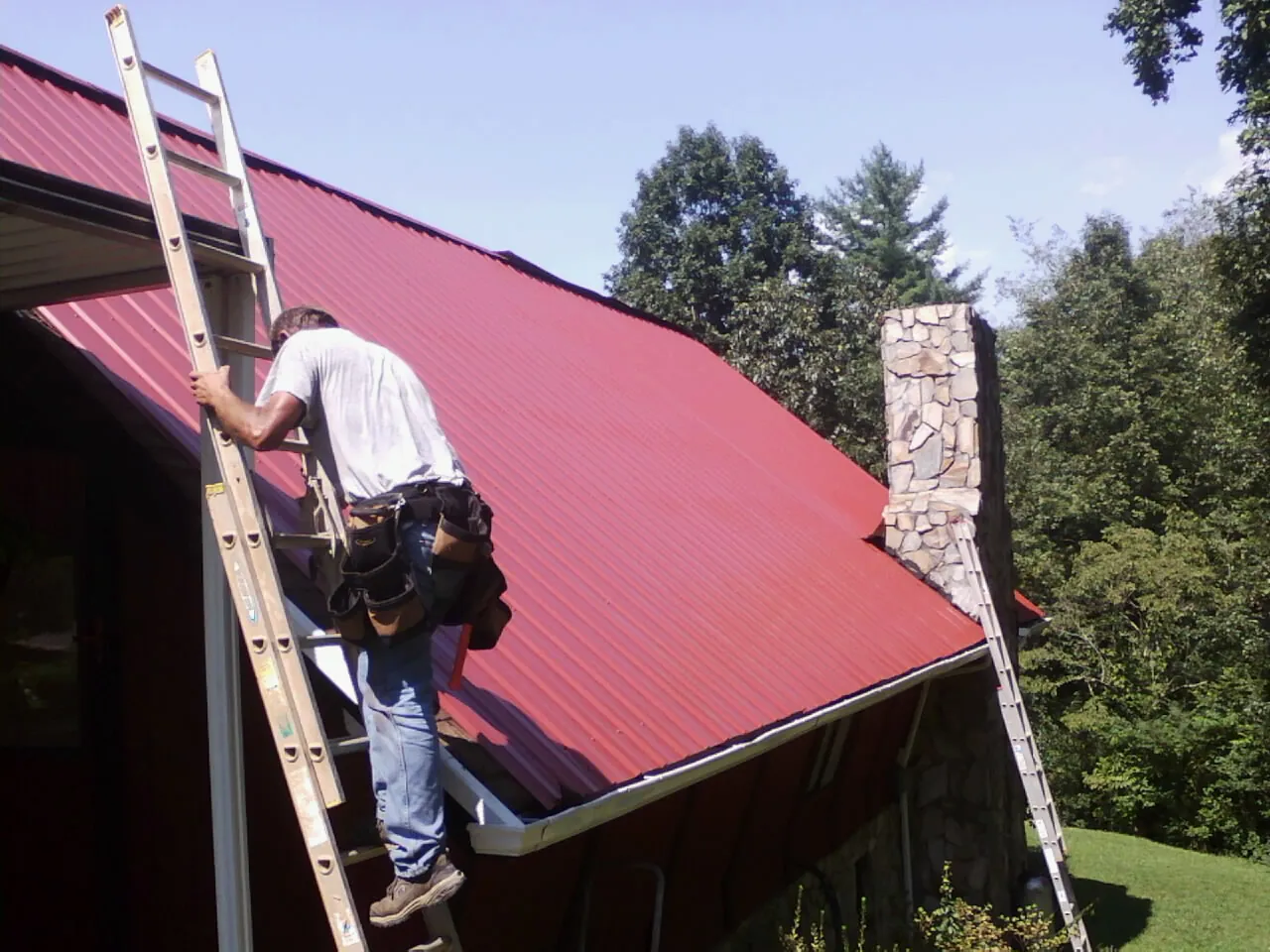 Roofer on a ladder beside a red metal roof and stone chimney — Soffit & Fascia Repair by Roofing Brothers in Marsteller